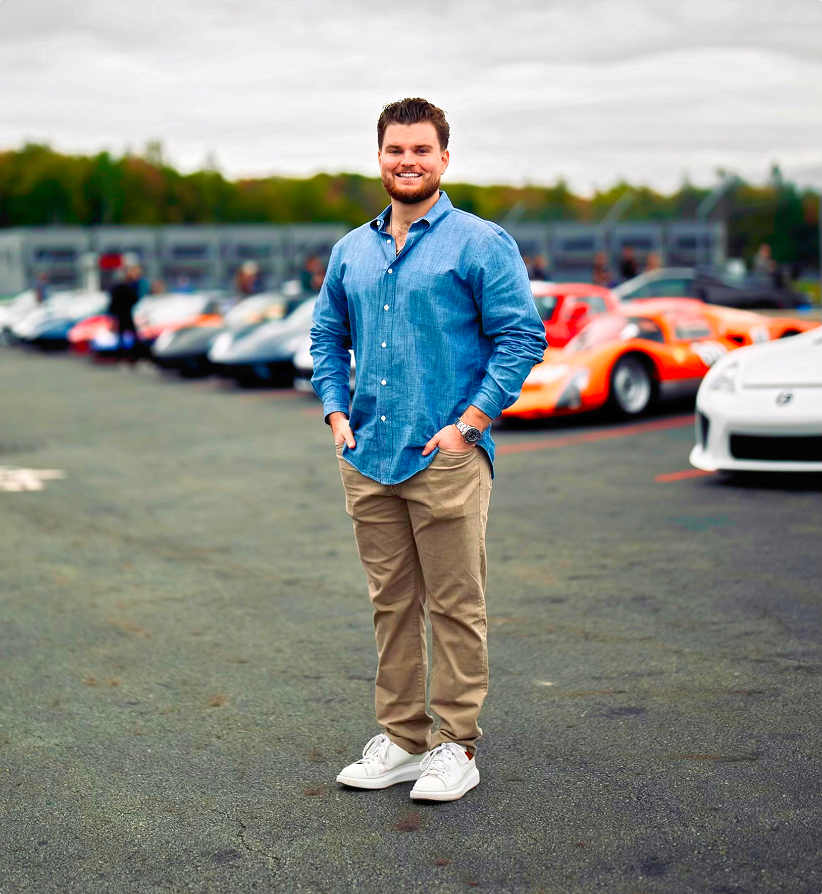 Photo of a man in a blue button down and tan pants with a row of diagonally parked sports cars in the background