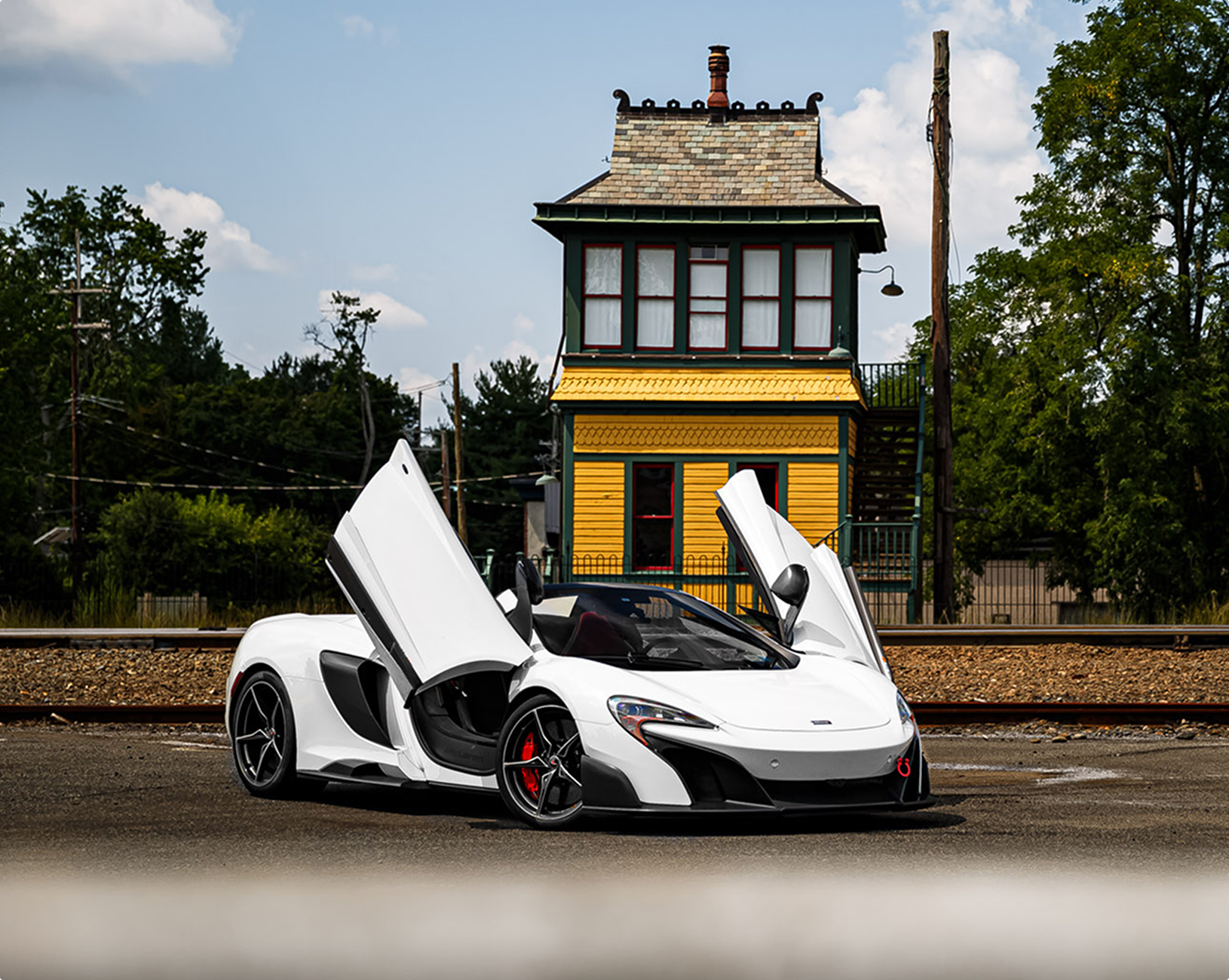 White McLaren 675LT Spider parked diagonally on asphalt with its doors up, train tracks and tower structure in the background.