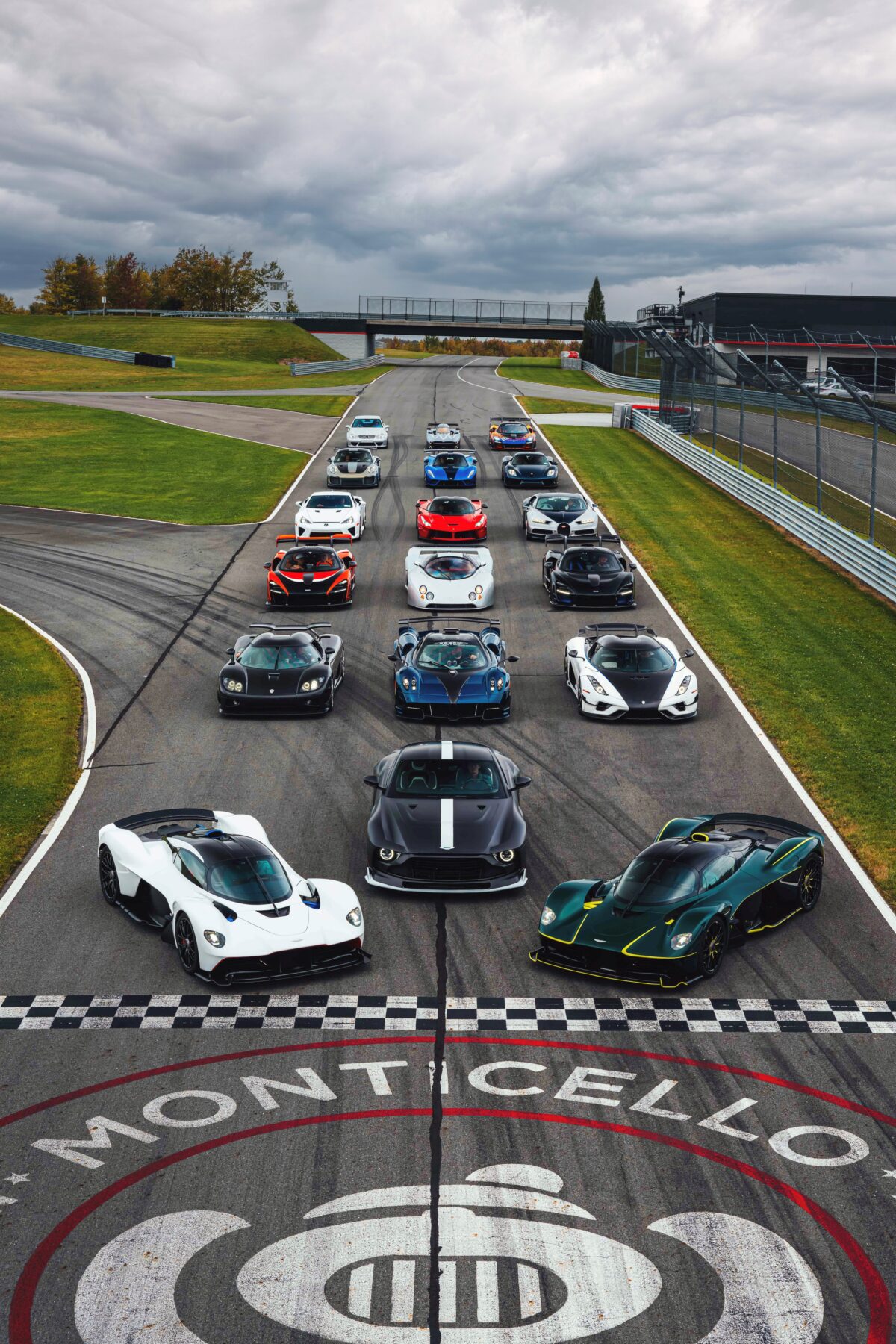 A lineup of hypercars and historic race cars staged on the Monticello Motor Club racetrack, gathered on the start-finish straight with the Monticello logo visible in the foreground.