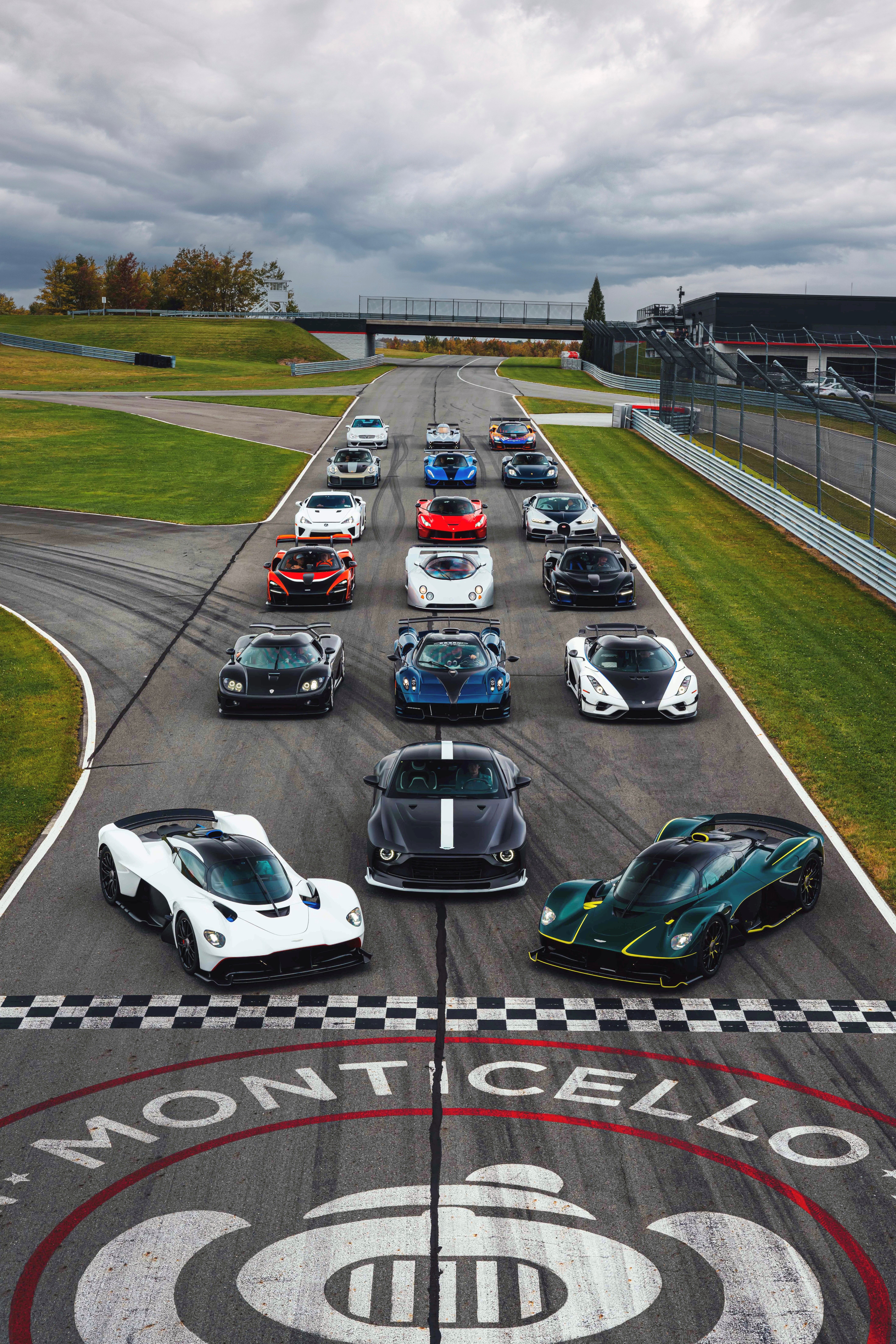 A lineup of hypercars and historic race cars staged on the Monticello Motor Club racetrack, gathered on the start-finish straight with the Monticello logo visible in the foreground.
